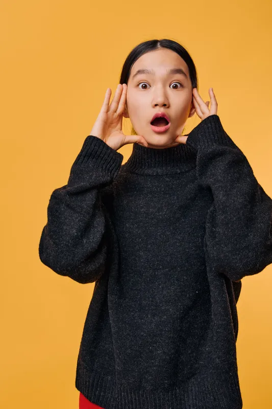 Young woman with surprised expression standing against orange background wearing black knitted sweater. Female showing shock and hands near face in casual fashion look with wide eyes and open mouth.