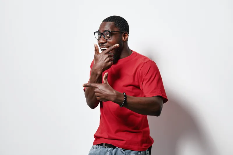 African American man with dark skin wearing glasses and red t shirt posing with hands framing face on white background. Casual style, smiling and confident young male portrait.