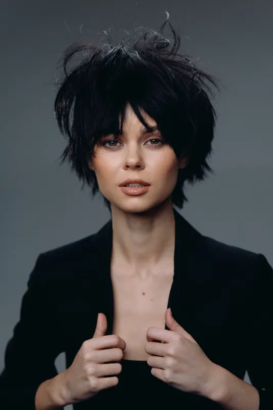 Portrait of a young woman with short black hair wearing a black jacket, serious expression on gray background.