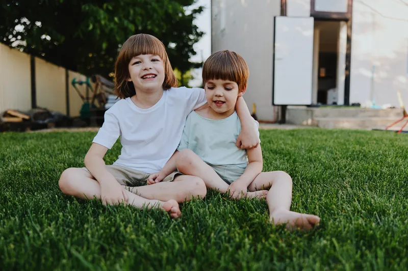 Happy boys sitting on green grass, enjoying a sunny day with smiles and playful expressions. A warm atmosphere and family-friendly vibes enhance the scene.