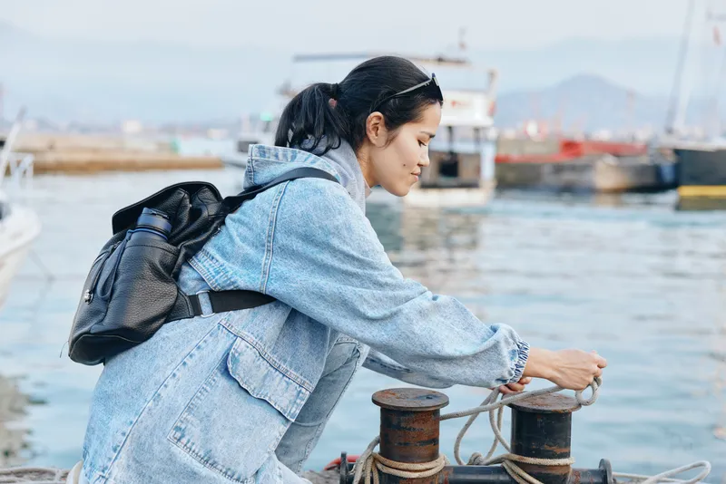 Woman in denim jacket at harbor docks handling rope and mooring, backpacked traveler beside the water, focusing on securing a boat line during outdoor maritime work