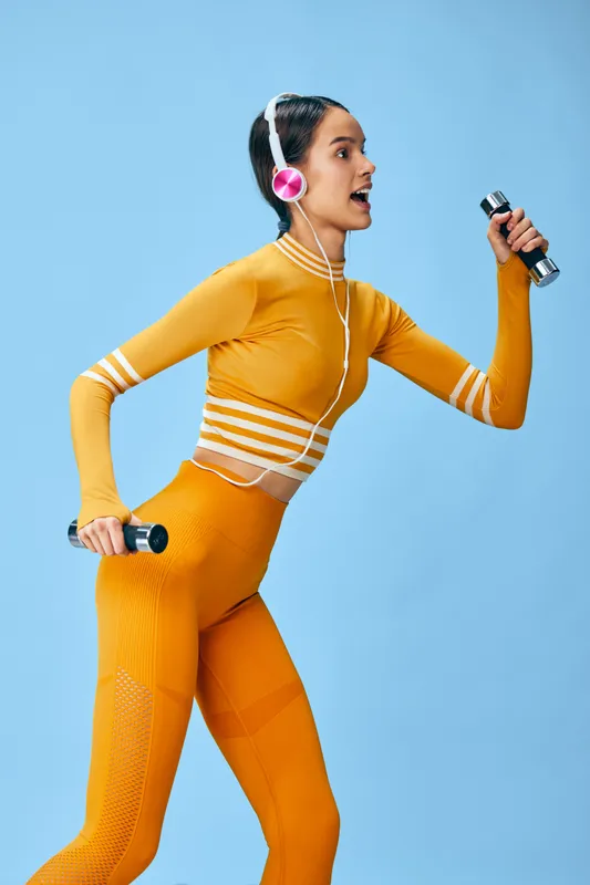 Vibrant fitness scene with a woman in bright orange activewear lifting dumbbells, wearing headphones, mid workout against a blue studio backdrop.