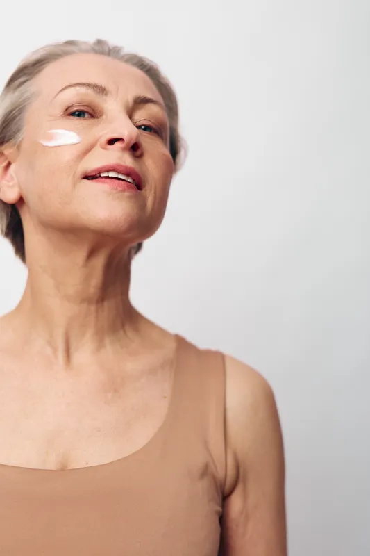 Mature woman with smooth skin applies white face cream on cheek, wearing beige tank top. Skincare concept for aging, beauty, wellness, and self-care on light background.