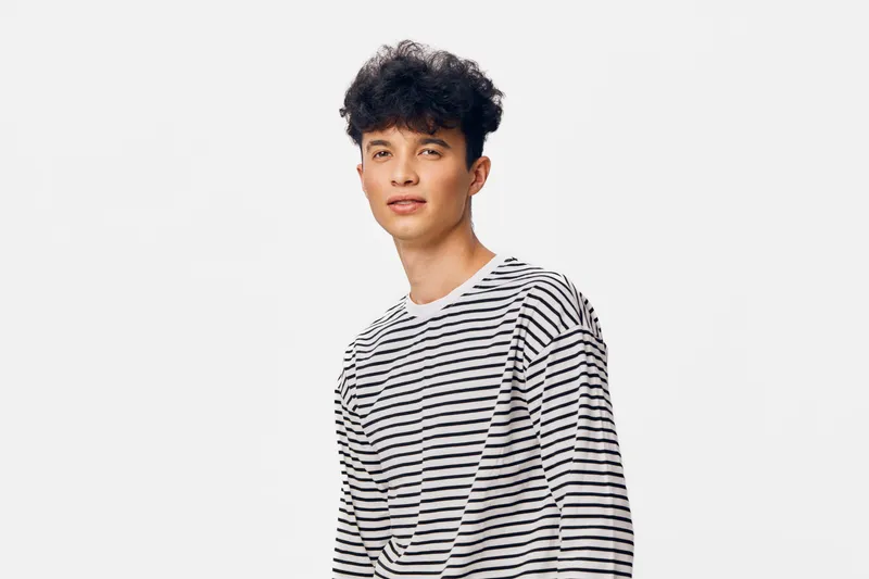 young man with curly hair wearing striped shirt stands confidently on bright plain background in casual outfit, natural expression, isolated portrait.