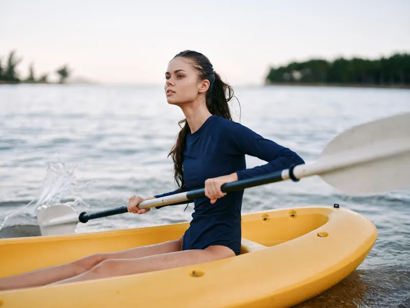 A young woman sits in a yellow kayak, paddling gently through calm water with an oar resting across her lap, focused on the horizon as soft light warms the scene.