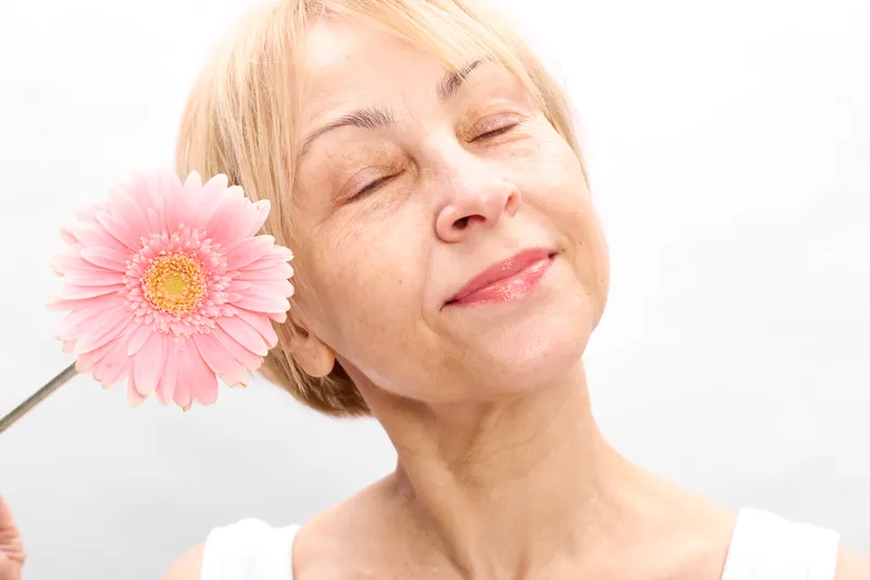Elderly woman holds gerbera flower closeup, eyes closed, smiling, beauty skincare concept, facial care, soft light, minimal white background, calm wellness mood