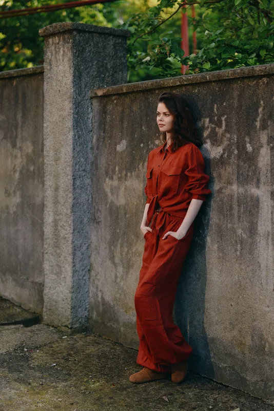 Portrait of a woman in a bold red jumpsuit leaning against a weathered wall in an outdoor urban setting, casual lifestyle mood, natural light, confident relaxed pose, street fashion
