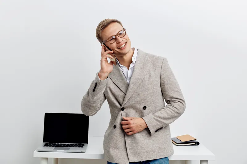 Smiling young professional man with glasses in stylish gray blazer talking on mobile phone while standing near modern workspace with laptop and notebooks isolated on plain white background. People lif