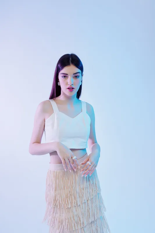 Young woman with long dark hair wearing a white crop top and fringed skirt poses against a soft blue background. Concept of beauty fashion style and modern elegance.