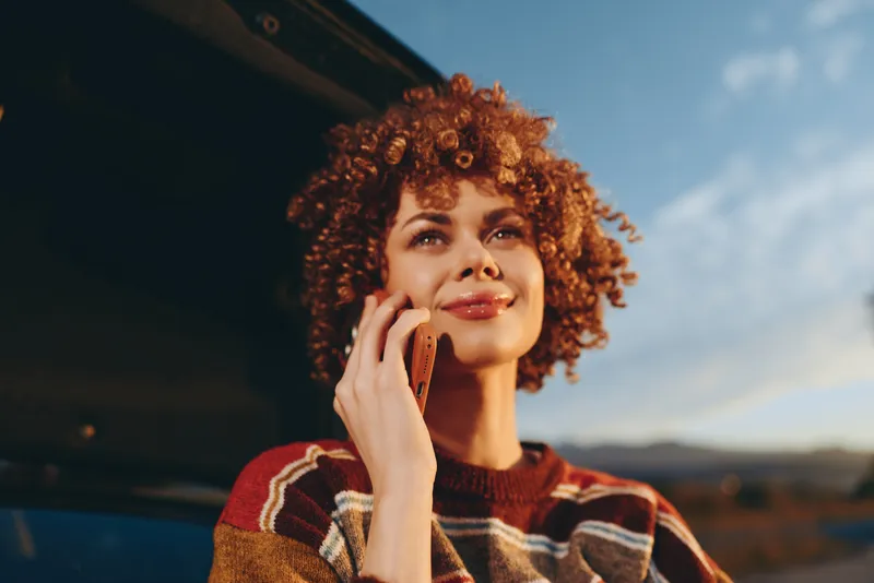 Woman lifestyle outdoors smiling curly phone call sunlight golden relaxed moment, cheerful eyes and warm sweater, hand holding phone near face, soft breeze, scenic sky and landscape background
