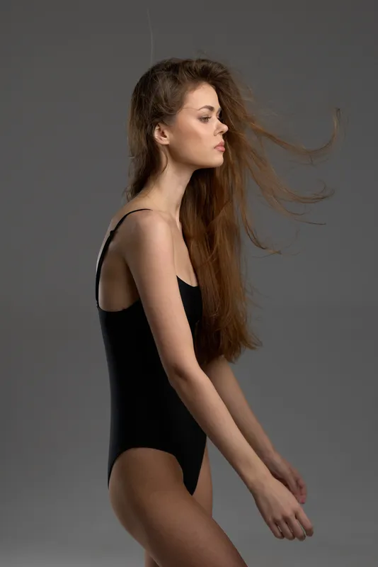 Profile of a young woman in a black swimsuit with windblown hair, posing against a grey background in a studio setting.