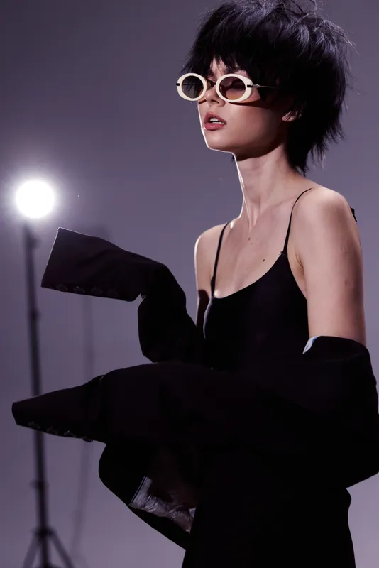 Stylish woman in black outfit wearing sunglasses with confident expression and modern hairstyle under studio lighting.