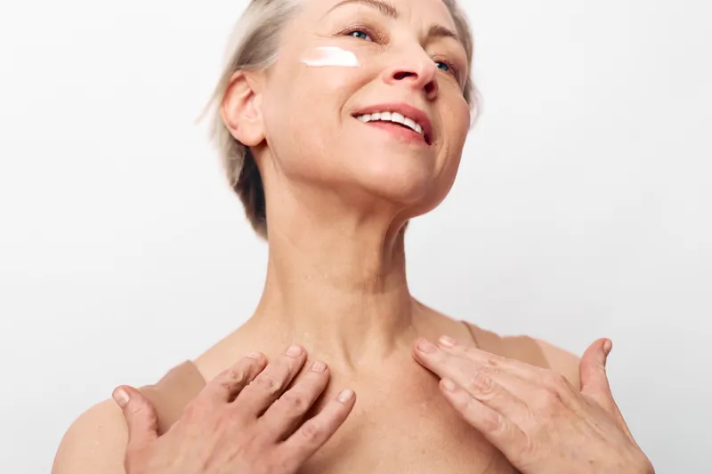 Mature woman with short hair applies cream on face and neck, smiling gently. Concept of skincare, antiaging, wellness, natural beauty, and selfcare in minimalistic studio lighting.