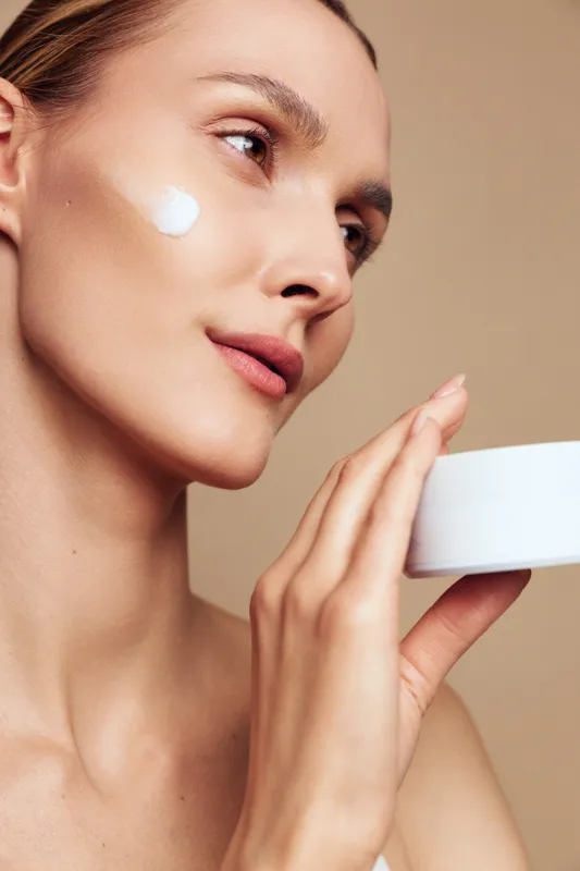 Young woman with clear skin applying white face cream on cheek holding open jar. Skincare routine concept with natural makeup and beige background. Soft lighting highlights healthy complexion.