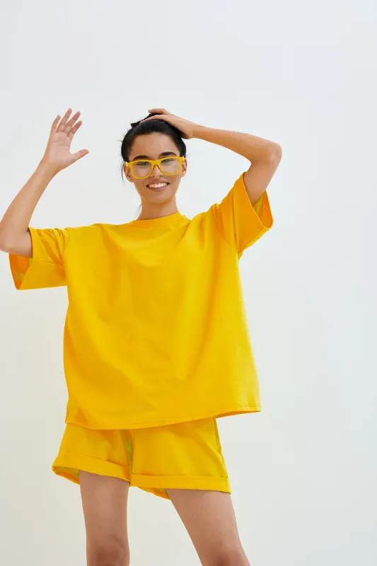 Young woman in bright yellow casual outfit and matching glasses smiles confidently against a minimal white background, showcasing modern summer fashion style.