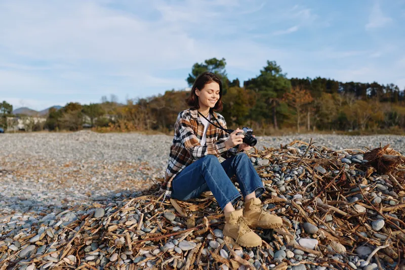 Woman photographer sits on a shell and driftwood beach, holding a camera, wearing a plaid shirt and boots, enjoying a sunny outdoor moment by calm water and autumn trees.
