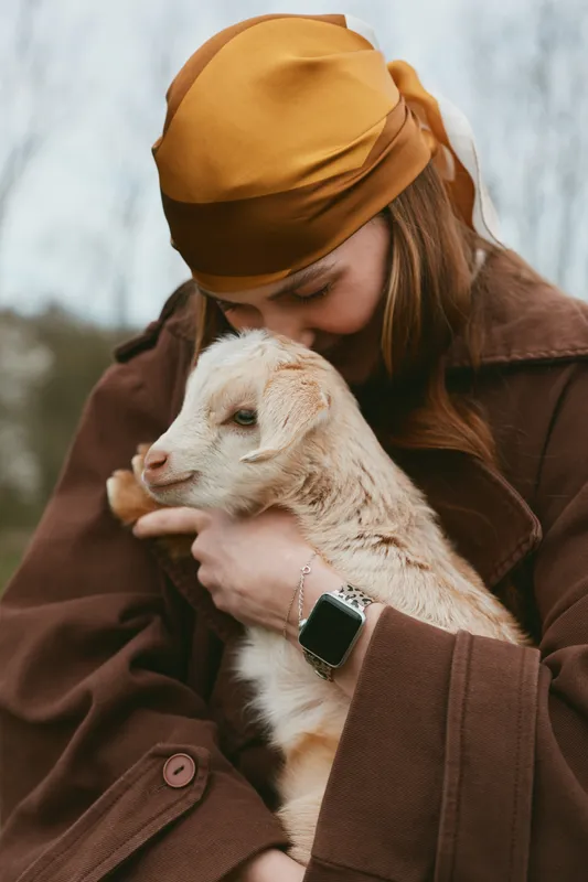 A woman holds a gentle goat kid in an outdoor autumn setting, capturing a lifestyle moment of affection and warmth, with rustic fashion and a cozy scarf in a natural farm environment.