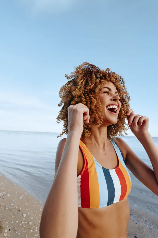 Woman beach sea summer swimsuit curly hair happiness laughter on the shoreline, she sings with fists raised, warm sunlight and blue sky, carefree vacation mood, outdoor relaxation