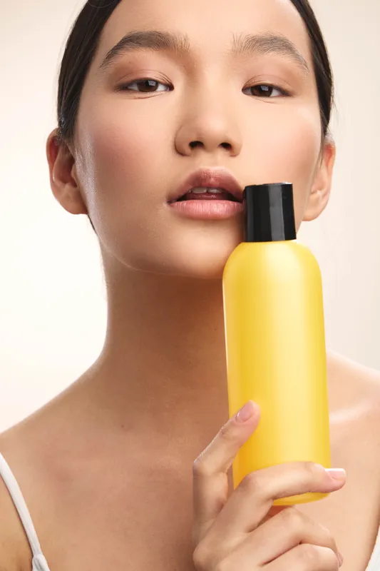 Asian woman gazes softly at the camera while lifting a bright yellow bottle, presenting a simple skincare moment in a clean studio setting, with a calm and confident expression.