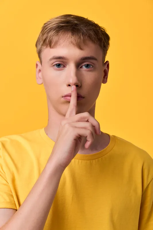 Hush gesture closeup of a youth in a yellow shirt, finger to lips signaling silence, capturing quiet focus, suspense, and a modern minimalist moment of secrecy