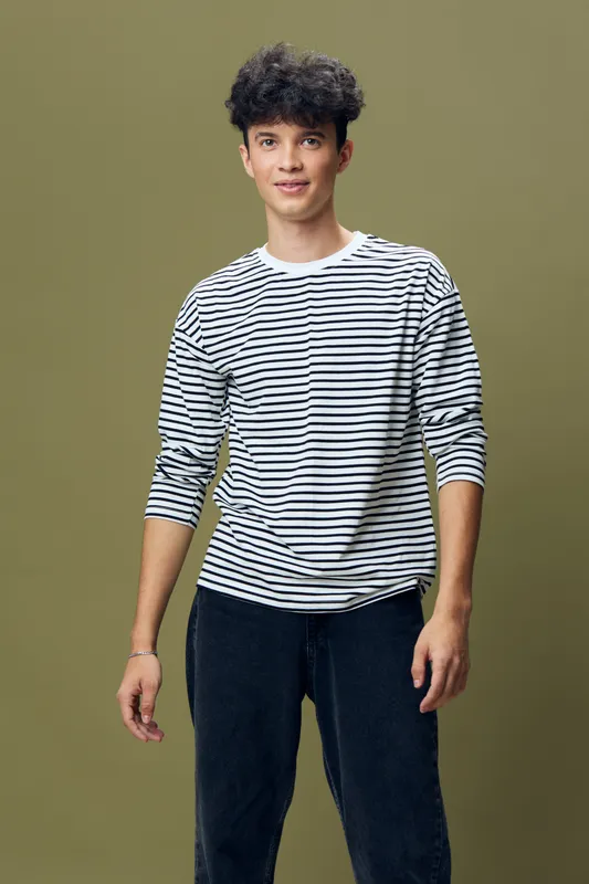young man standing wearing a black and white striped long sleeve shirt with black pants, casual style, curly hair, olive background, relaxed expression, modern fashion