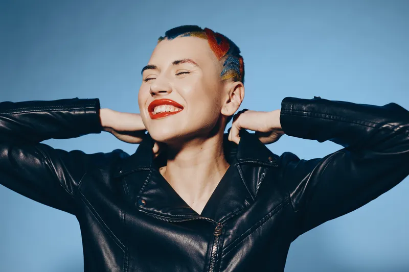 Smiling woman with colorful short hair wearing a black leather jacket, expressing joy and confidence against a solid blue background in a modern studio portrait.