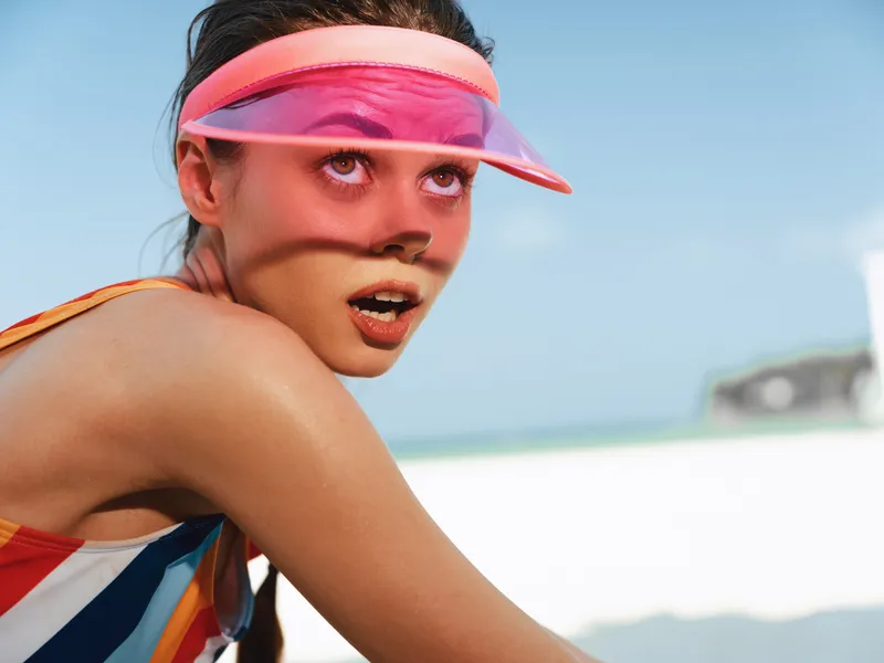 Close-up of a young woman wearing a pink visor at a sunny beach, displaying a surprised expression with sunlit skin, vibrant striped clothing, and a blurred ocean backdrop.