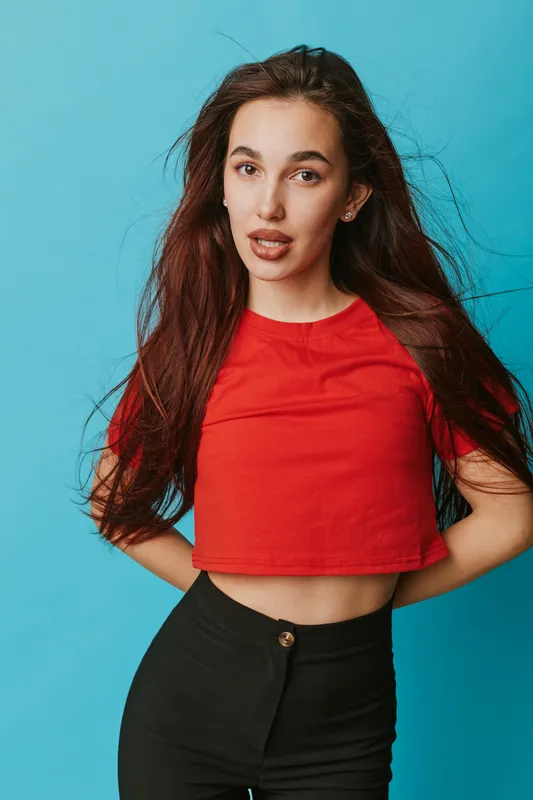 Young woman with long hair wearing red crop top and black pants posing with hands behind back on blue background in studio. Fashion, casual style and confident expression portrait.
