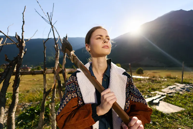 Woman, axe, outdoors, mountains, countryside, wood — young woman holding a chopping axe in a rural field at sunrise, rustic portrait with fence, cozy jacket, calm expression and woodcutting chore.