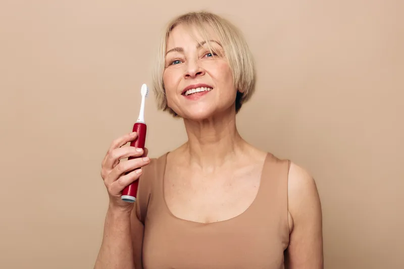 Smiling mature woman with short blonde hair holding electric toothbrush, standing against beige background. Concept for dental care, oral hygiene, and healthy lifestyle habits.