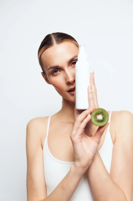 Smiling woman with light makeup holds a white bottle near her face, a kiwi slice adds a natural touch. Close up on skincare, beauty routine, healthy glow, fresh and clean.