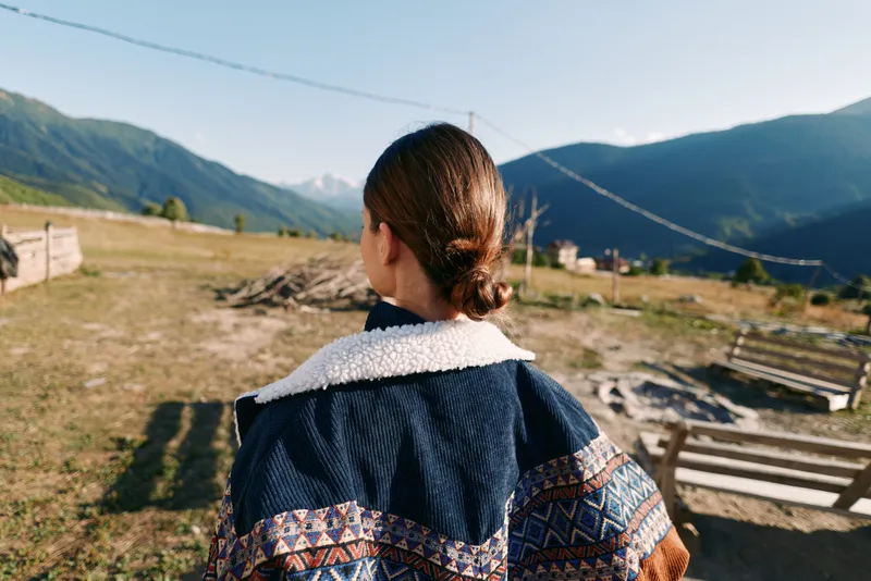 Woman back view in rural countryside wearing patterned jacket, overlooking mountain landscape and valley. Outdoor travel scene with natural light, calm morning and wooden fence.