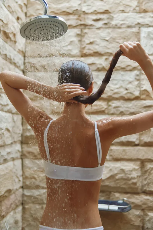 woman showering outdoors with water falling, holding hair, stone wall background, natural light, summer wellness moment, back view of adult female.