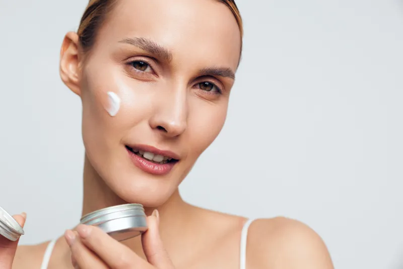 Close-up of a smiling woman applying white face cream on her cheek. Concept of skincare, beauty, and self-care with soft lighting on a neutral background. Healthy skin routine.