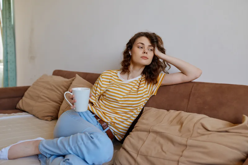 Calm woman lounging on a sofa at home, striped yellow shirt, blue jeans, holding a mug, soft natural light, creating a cozy lifestyle moment focused on relaxation, warmth, and everyday comfort.