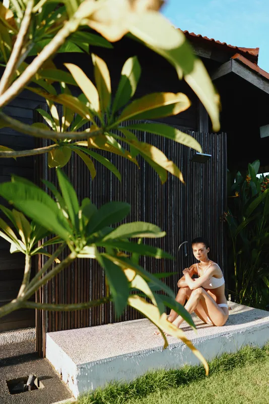 Young woman sitting on a low concrete ledge beside a dark wooden wall, enjoying outdoor calm in bright sun and fresh air, relaxed and thoughtful in a private moment
