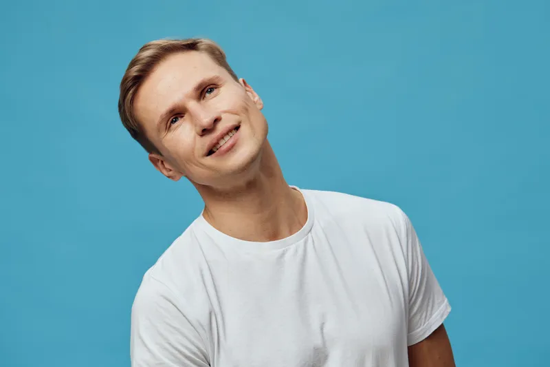 Happy smiling young man with light brown hair wearing a white t-shirt is looking at the camera against a plain blue background. Casual pose, positive mood, studio shot, people lifestyle concept.