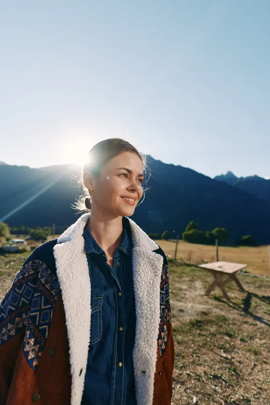 Woman portrait in mountains at sunrise with sunlight flare, smile and warm jacket over denim shirt. Outdoors field scene with calm rural landscape and soft backlight.