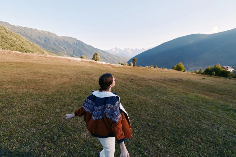 Woman walking in a meadow toward mountains across open landscape, nature hiking outdoors travel scene with jacket and shawl, back view capturing freedom and scenic valley peace.