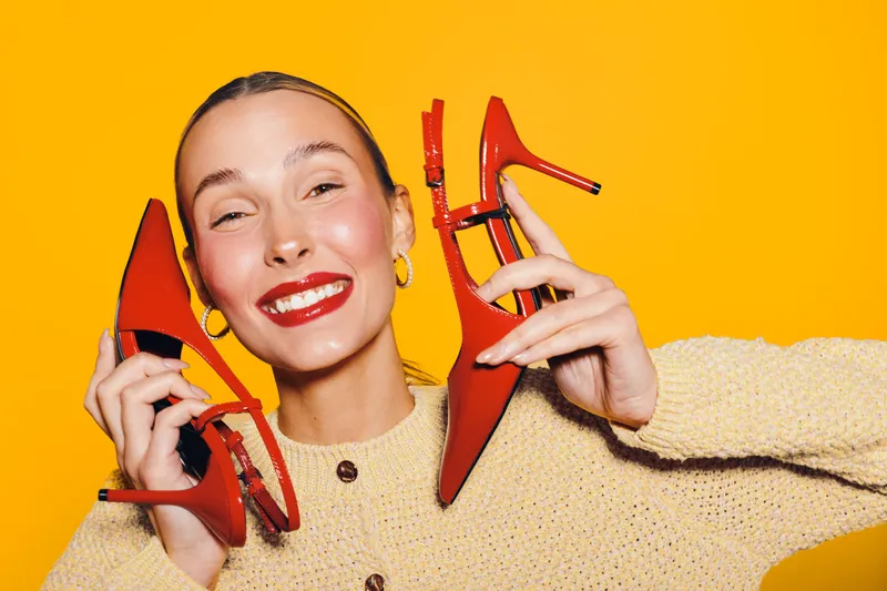 Happy woman with red lipstick holds red high heel shoes near face on yellow background. Bright studio lighting highlights joyful expression and fashion concept with vibrant colors.