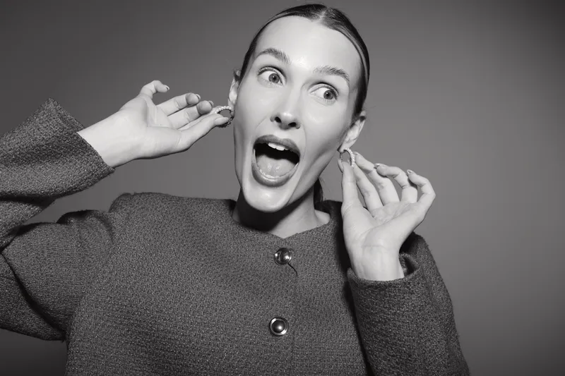 Surprised Caucasian woman with light skin holds earrings near ears, mouth open in shock, wearing textured jacket in studio. Concept for fashion, emotions, jewelry, and beauty.