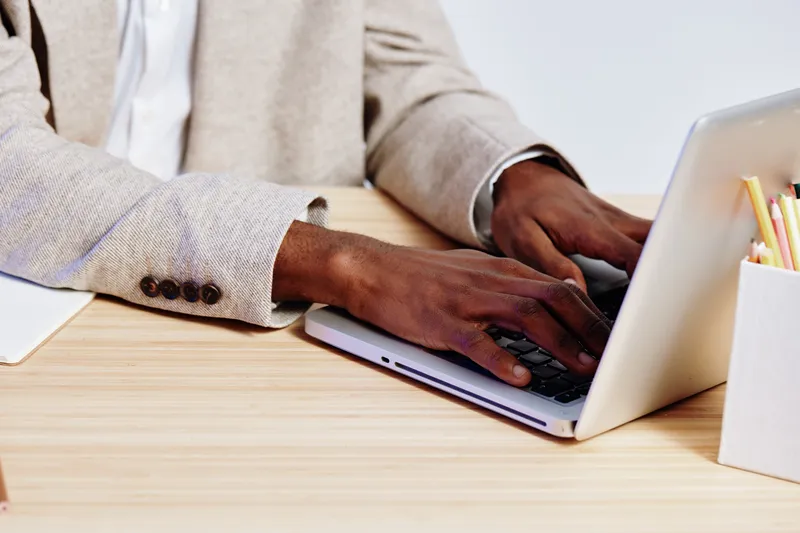 Close-up of man’s hands typing on a silver laptop at a wooden desk, wearing a light beige blazer and white shirt, with notepad and colorful pencils nearby. Focus on productive work environment, office