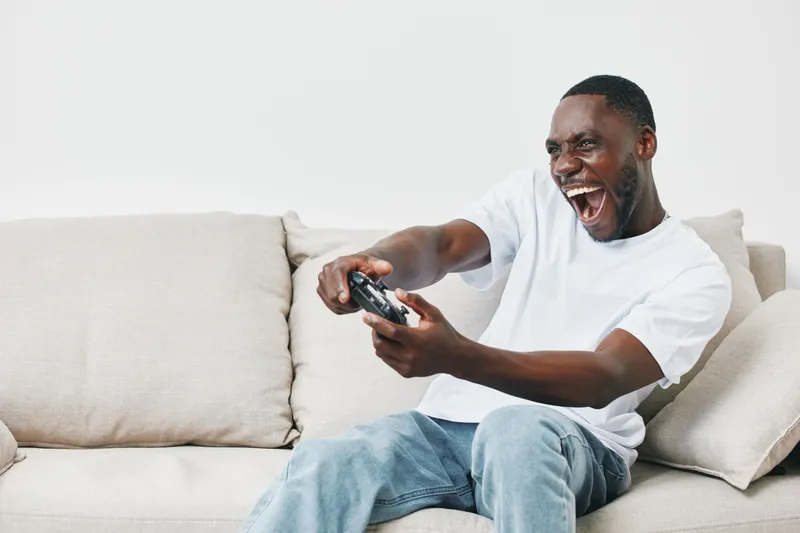 Excited young man playing video games on a couch, expressing joy and enthusiasm, wearing casual clothing in a modern living room setting