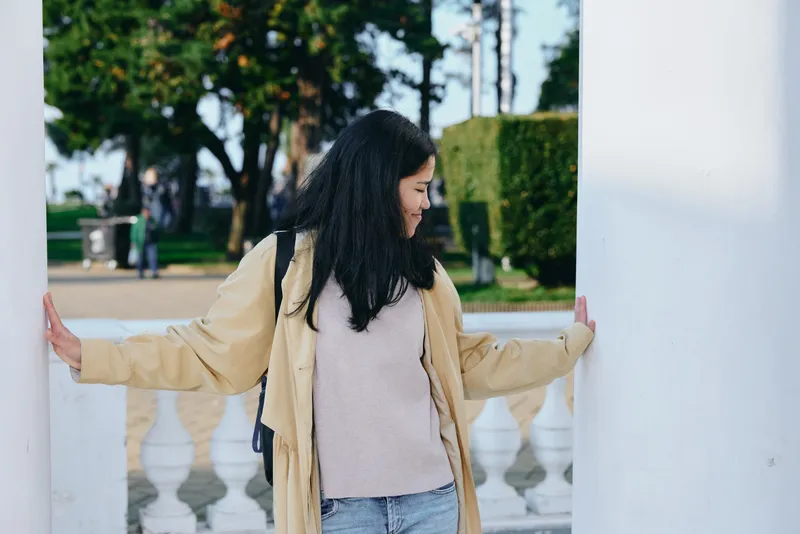 Woman in a casual coat and sweater stands between white pillars in a park, outdoors, posing with calm, candid mood, fashion style and architectural backdrop, natural light and soft colors creating a