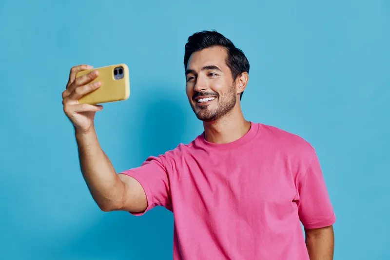 Happy smiling man in bright pink t-shirt taking selfie with smartphone on solid blue background. Modern lifestyle and social media engagement concept.