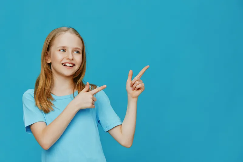 Happy girl in a blue t shirt pointing to the side against a blue background, expressing joy and enthusiasm while promoting positivity