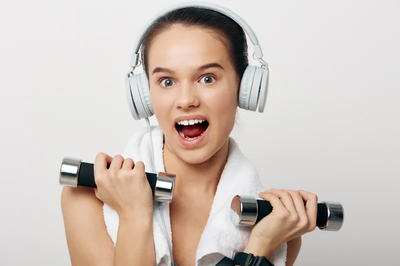Fitness woman lifting dumbbells with headphones, energetic workout scene in a studio, towel around shoulders, showcasing strength, motivation and a healthy lifestyle.