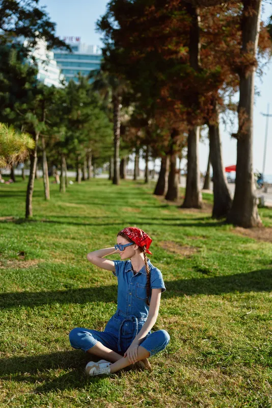 lifestyle woman in modern boho-western denim street style sits on grass outdoors with sunglasses and red bandana, film color tone captures relaxed casual mood