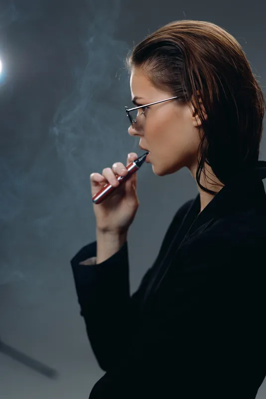 Young woman smoking electronic cigarette vaping indoors with trendy glasses and dark outfit, exhaling vapor against dark background.