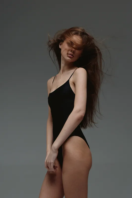 slender woman in black bodysuit with tousled hair posing against gray background, elegant and modern style